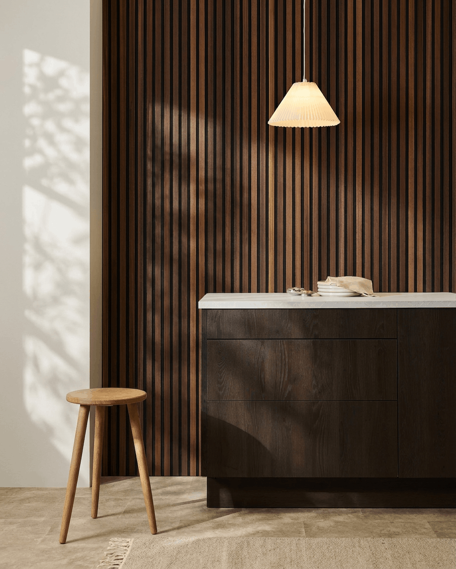 Minimalistisches Interieur mit einem Holzhocker, einem Schrank aus dunklem Holz, einer Pendelleuchte und einem WoodUpp Akupanel Akustikpaneel aus Holzlamellen in Natural Wood/Räucherlärche.